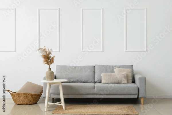 Fototapeta Stylish sofa, basket with pillow and table near white wall