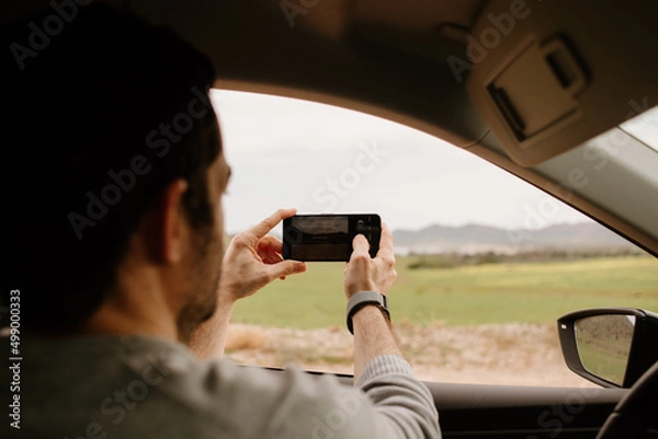 Obraz On a road trip. Young man stops the car to take a photo of the landscape sunset.