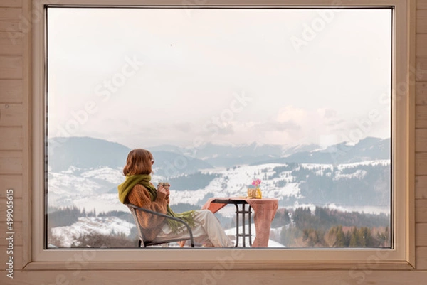 Fototapeta Woman sitting with a drink on terrace of tiny house in the mountains, view through the window from the inside. Concept of small modern cabins for rest and escape to nature