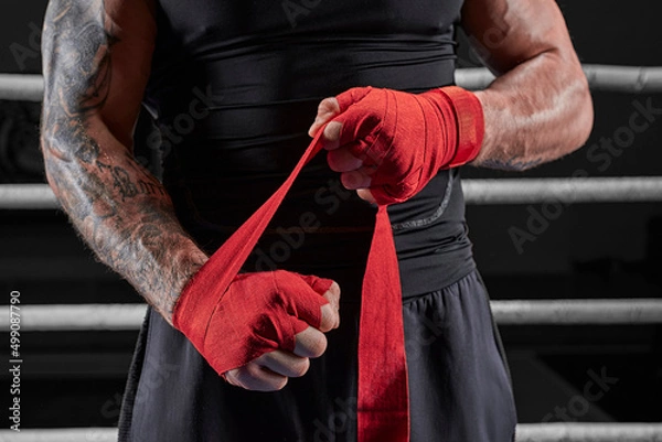 Obraz Red bandages on the hands of a kickboxer against the background of the ropes of the ring. The concept of mixed martial arts.