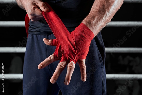 Obraz Red bandages on the hands of a kickboxer against the background of the ropes of the ring. The concept of mixed martial arts.