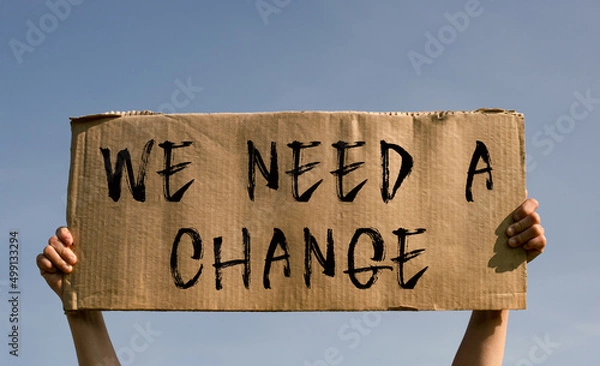 Fototapeta A poster with the words WE NEED A CHANGE in the hands of a young girl against a blue sky.
