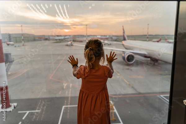 Fototapeta Little girl at the airport is waiting for landing at the big window. Cute kid stands at the window against the backdrop of airplanes. Looking forward to leaving for a family summer vacation