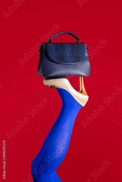 Fototapeta A blue foot in a beige shoe with high heels is raised up on a red background. Women's legs upside down with a blue handbag on high-heeled shoes.