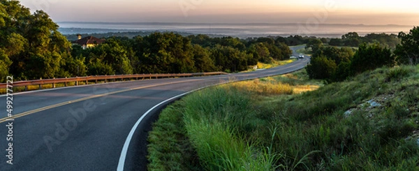 Fototapeta S shape curved road winding through a hilly terrain with a guard rail and a vehicle and hills in the distance, Guadalupe Valley, Texas