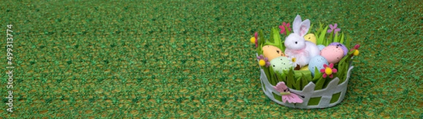 Fototapeta A small white rabbit sits in a basket with small multi-colored eggs on a green background, banner size.