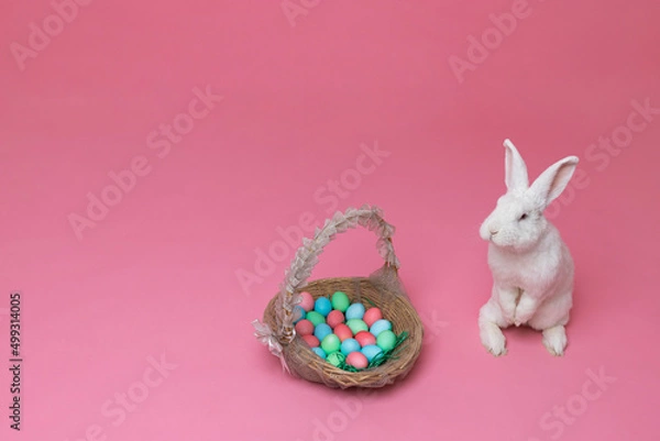 Fototapeta A white rabbit stands on its hind legs on a pink background. An Easter Bunny is sitting next to a basket in which there are multi-colored painted chicken eggs.