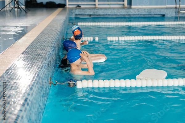 Obraz A group of boys and girls train and learn to swim in a modern swimming pool with an instructor. Development of children's sports. Healthy parenting and promotion of children's sports.
