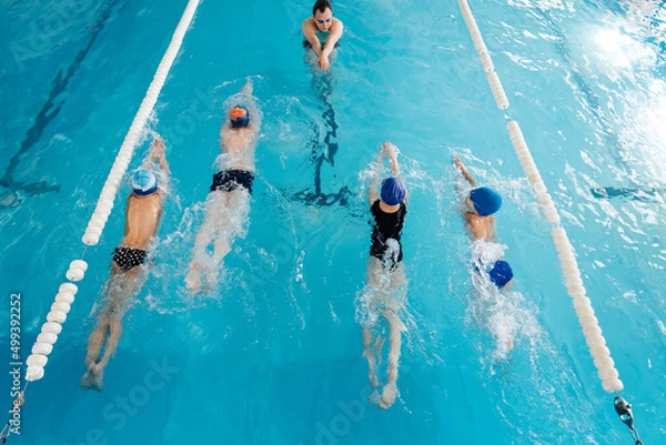 Fototapeta A group of boys and girls train and learn to swim in a modern swimming pool with an instructor. Development of children's sports. Healthy parenting and promotion of children's sports.