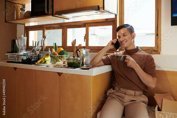 Fototapeta Smiling young man sitting at kitchen counter, drinking morning coffee and talking on phone with friend