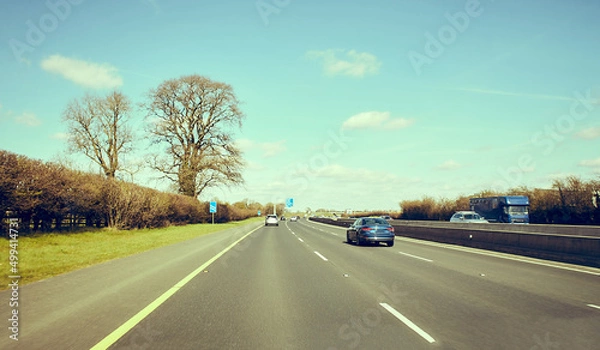 Obraz Rear view of cars driving on motorway, Ireland. Road with metal safety barrier or rail. cars on the asphalt under the cloudy blue sky. Highway traffic.