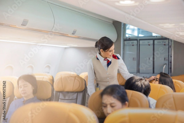 Obraz Beautiful female flight attendants checking the passengers on safety standard, seat belt, turn off electric devices, seat back and tray table in upright position before airplane take off and landing.