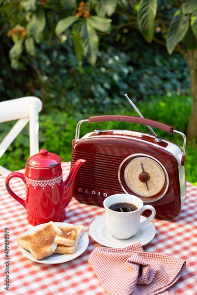 Obraz A white cup with coffee, red coffee pot and radio are on a table in a summer garden. Outdoor breakfast theme. A vertical image.