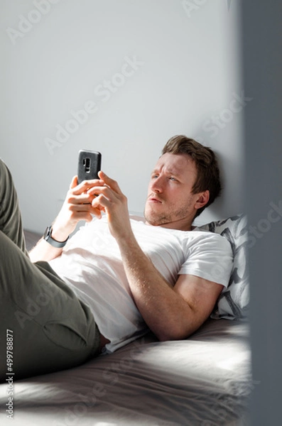 Fototapeta Caucasian attractive young man in white t-shirt woke up chatting, surfing, working on pc and making phone call on bed in bedroom interior. Modern device for remote communication