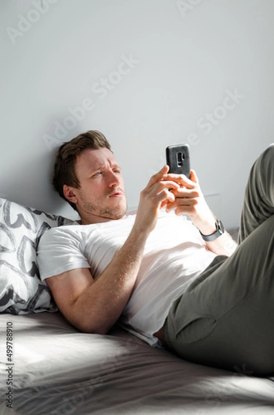 Fototapeta Caucasian attractive young man in white t-shirt woke up chatting, surfing, working on pc and making phone call on bed in bedroom interior. Modern device for remote communication