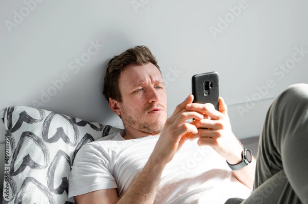 Fototapeta Caucasian attractive young man in white t-shirt woke up chatting, surfing, working on pc and making phone call on bed in bedroom interior. Modern device for remote communication