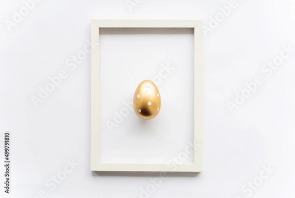 Fototapeta Golden egg with polka dots on a white background with copy space for text in a frame. Minimal concept. View from above. Easter card, soft selective focus.