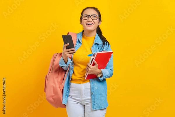 Fototapeta Portrait of smiling young Asian woman student in denim clothes, glasses with backpack holding mobile phone and books isolated on yellow background. Education in high school university college concept