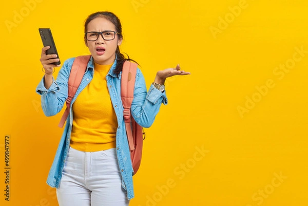 Fototapeta Portrait of  confused young Asian woman student in casual clothes with backpack holding mobile phone and saying no with palms isolated on yellow background.  Education in college university concept