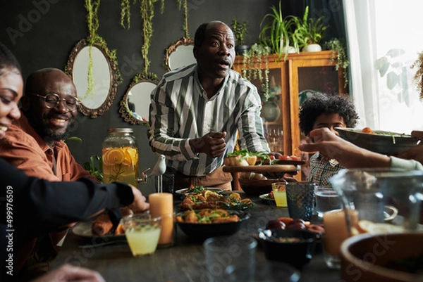 Fototapeta African mature man suggesting dish for his family while they sitting at table in dining room