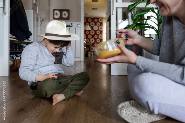 Fototapeta Mother feeding a boy with autism wearing a hat, sitting on the floor while playing with the mobile phone