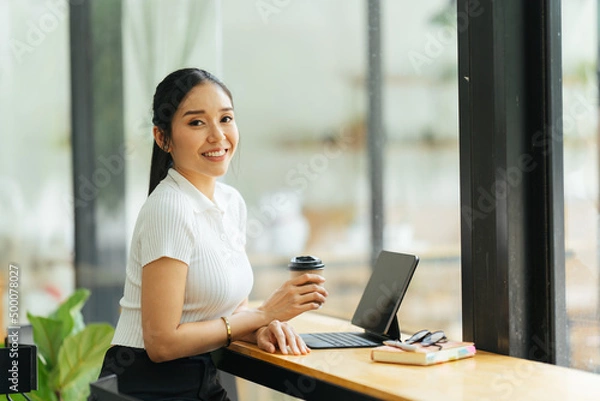 Obraz Beautiful young asian business woman drinking coffee and using laptop computer while working in office.