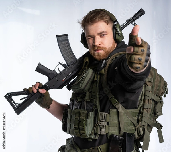 Fototapeta the airsoft player puts his rifle on his shoulder and aims at us with his fingers. a man in an outfit, in headphones, a bulletproof vest, with a backpack on a white background.