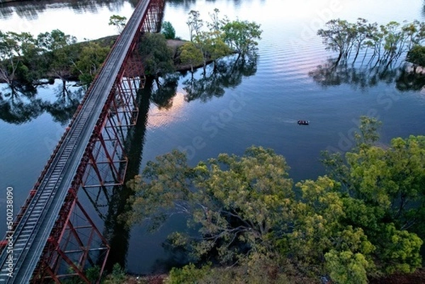 Obraz drone shot of a boat at sunset on a river with a train bridge