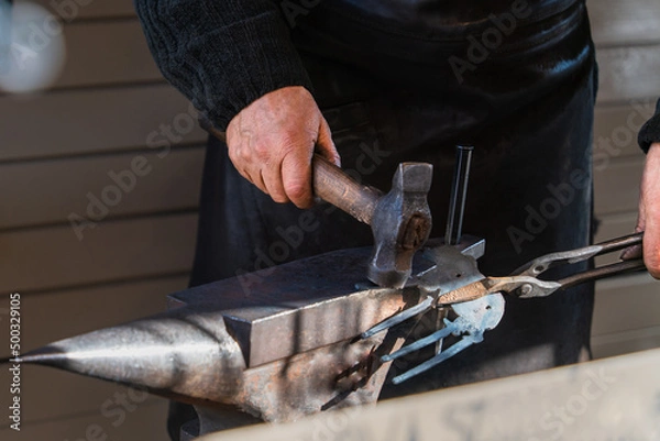 Obraz Selective focus close-up of a blacksmith's hand with a hammer on a metal anvil and a metal part, a beam of sunlight illuminates the metal. Illustration of hard manual labor and craftsmanship