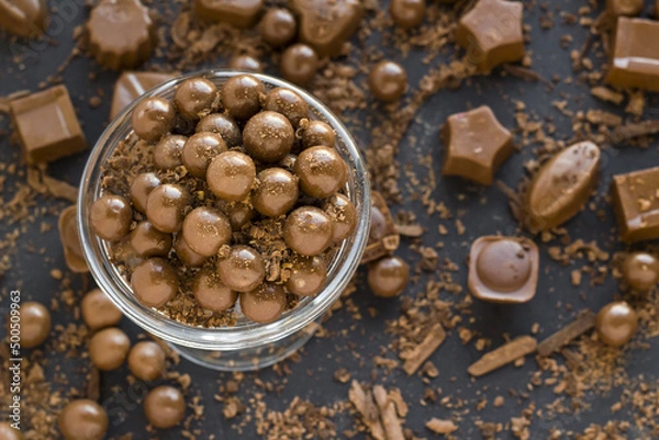 Fototapeta Milk chocolate covered hazelnuts with grated chocolate chips in an elegant bowl, with shaped chocolates on black surface,
