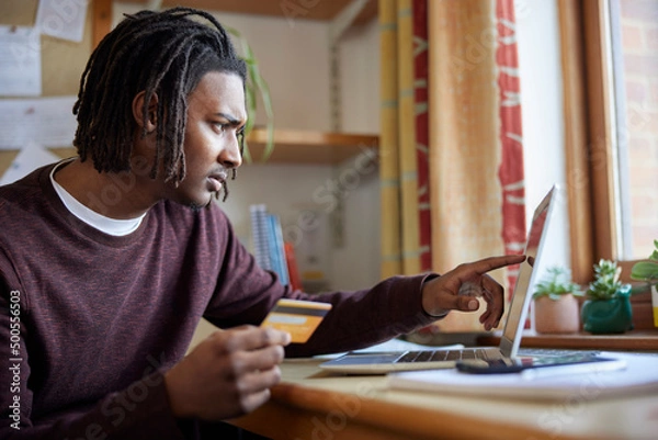 Obraz Male University Or College Student With Credit Card Looking At Laptop Worried About Debt At Desk In Room