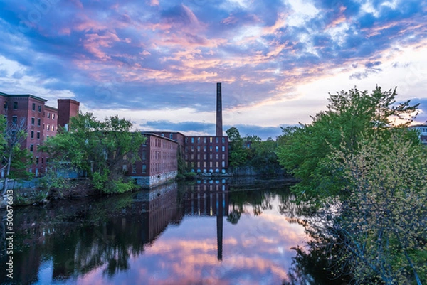 Fototapeta Historic cotton mill building with a tall brick chimney on the banks of the river in the old Nashua Industrial Park and its reflection in the Nashua River. New Hampshire, USA