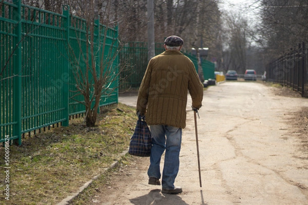 Obraz Pensioner in Russia. Poverty in world. Elderly man walks leaning on walking stick. Background man goes into distance.
