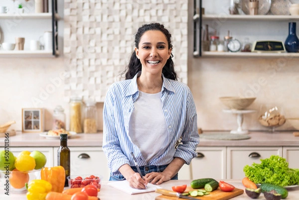 Obraz Happy housewife writing new food recipes in her notebook, standing in home kitchen interior and smiling at camera