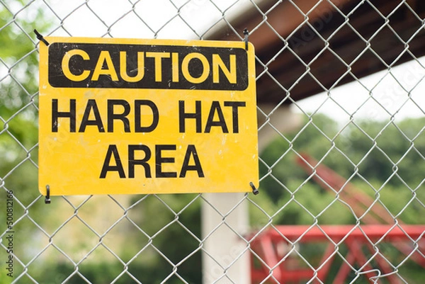 Fototapeta Yellow "caution hard hat area" sign on a chain link fence with a newly constructed bridge and crane in the background.