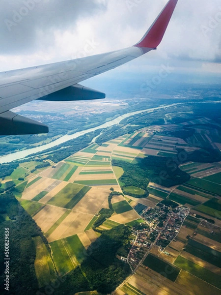 Fototapeta Beautiful view from the window on the wing of the aircraft and the multi-colored landscape of Austria. The plane flies below the clouds and prepares to land in Vienna. Vertical. Vienna, Austria