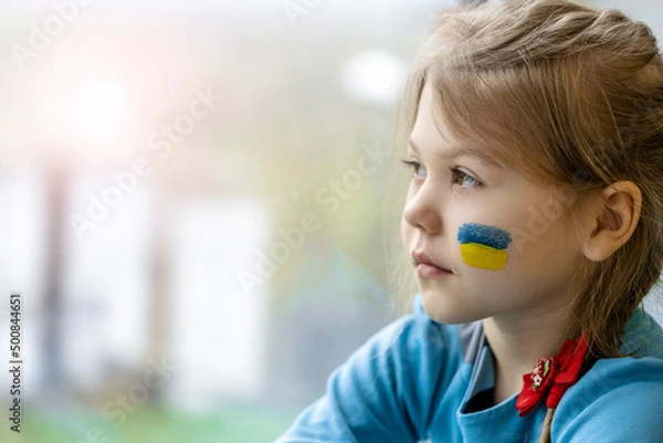 Fototapeta Portrait of an 8-year-old girl with a Ukrainian flag on her face, sitting by the window.