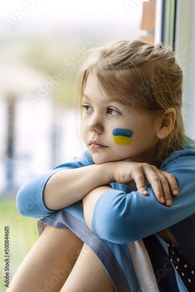 Fototapeta Portrait of an 8-year-old girl with a Ukrainian flag on her face, sitting by the window.