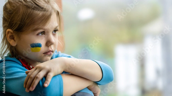 Fototapeta Portrait of an 8-year-old girl with a Ukrainian flag on her face, sitting by the window.