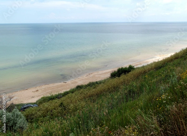 Fototapeta Azov sea coastal area with smooth green grass slope. Seashore view with sandy undeclared beach, blue sky horizon and a car.