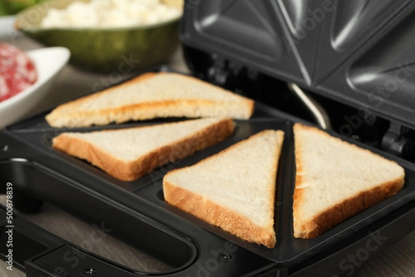 Fototapeta Modern sandwich maker with bread slices on wooden table, closeup