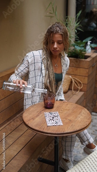 Fototapeta Portrait of an attractive girl with dreadlocks and boho style jewelry with a bottle of water in a cafe. Hippie girl sits in a cafe, drinks water and smiles. Close portrait, bohemian style