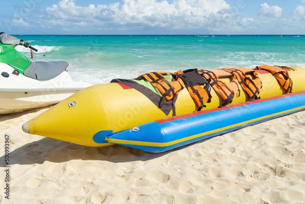 Fototapeta Inflatable bananas for group skating on the sea. Active rest,  attraction on the water. Banana boat with life jackets on a sandy beach against the backdrop of the sea.
