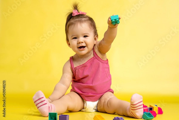 Fototapeta A 1 years old girl is playing with a children's multi-colored constructor. The girl is sitting on a yellow background in a diaper.