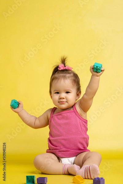 Fototapeta A 1 years old girl is playing with a children's multi-colored constructor. The girl is sitting on a yellow background in a diaper.