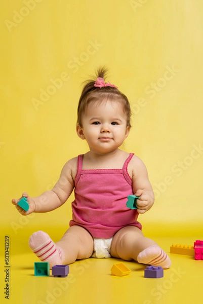 Fototapeta A 1 years old girl is playing with a children's multi-colored constructor. The girl is sitting on a yellow background in a diaper.