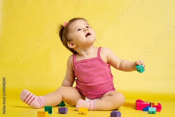 Fototapeta A 1 years old girl is playing with a children's multi-colored constructor. The girl is sitting on a yellow background in a diaper.