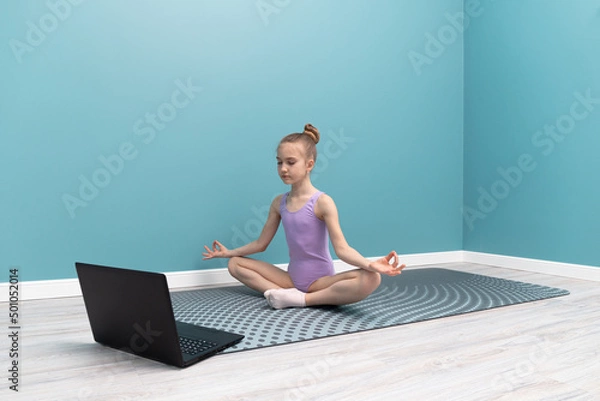 Fototapeta A young girl is doing gymnastics in front of a laptop. Classes at home