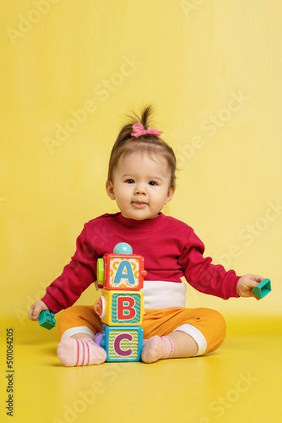 Fototapeta Girl 1 year old, playing with educational cubes with the English alphabet. The girl is sitting on the floor on a yellow background.