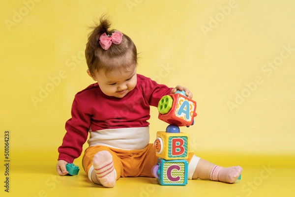 Fototapeta Girl 1 year old, playing with educational cubes with the English alphabet. The girl is sitting on the floor on a yellow background.